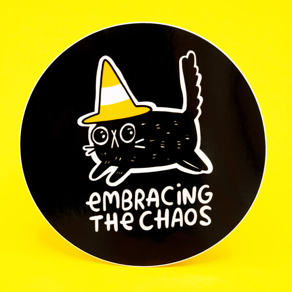 A round black vinyl sticker by Katie Abey featuring a quirky black cat wearing a yellow and white traffic cone hat with the words “Embracing The Chaos” below. Printed in bold white line art with a playful, weird vibe. Premium durable vinyl, 10cm size, perfect for laptops, notebooks, and water bottles. Bright yellow background.