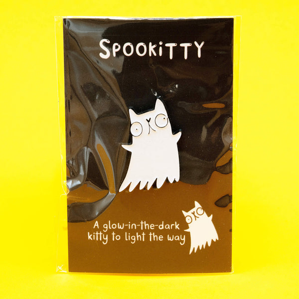 A Katie Abey Spookitty glow in the dark glitter enamel pin badge shown on its black backing card inside clear packaging against a bright yellow background. The ghost cat enamel pin has a white glitter finish and a bold black outline. Fun spooky themed accessory for bags, denim jackets, and pin collections.