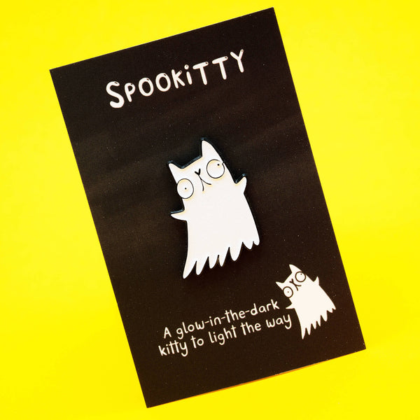 A Katie Abey Spookitty glow in the dark glitter enamel pin badge displayed on a black backing card against a bright yellow background. The white ghost cat design features big round eyes, a black outline, and a wavy ghost tail. Cute spooky themed enamel pin perfect for jackets, bags, and accessories.
