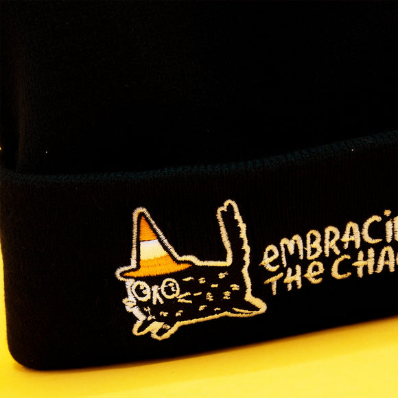 Close up of the embroidered design on the black Embracing The Chaos Cat Beanie Hat by Katie Abey. The detail shows a cute chaotic cat wearing a candy corn coloured hat with the words Embracing The Chaos. Ethically made quirky beanie perfect for alternative fashion and Katie Abey fans.