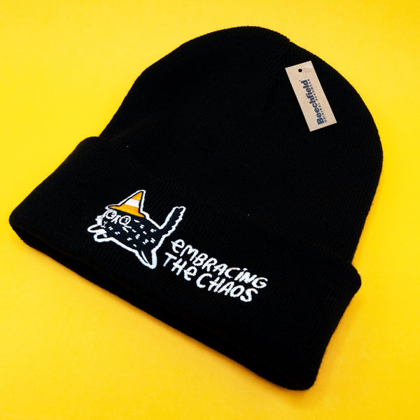 A black Embracing The Chaos Cat Beanie Hat by Katie Abey shown on a bright yellow background. The beanie features an embroidered quirky black cat wearing a candy corn coloured hat with the text Embracing The Chaos stitched in white. Cosy ethically made winter hat perfect for quirky fashion lovers and fans of Katie Abey.
