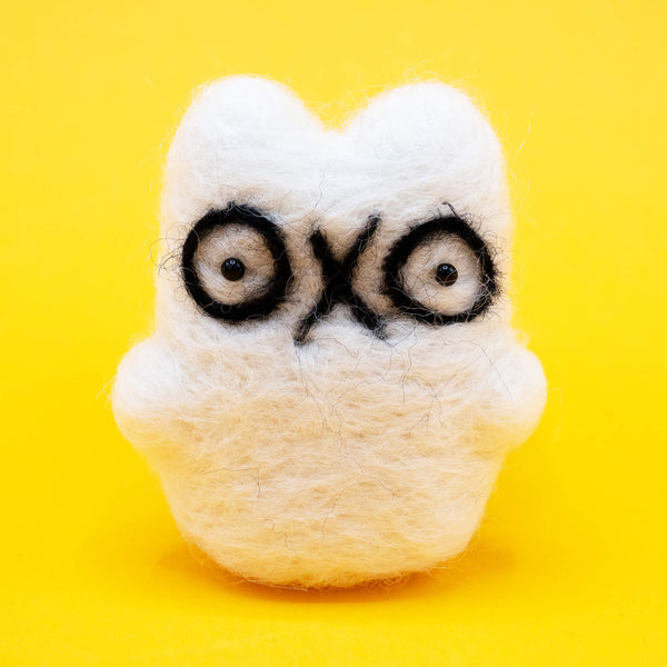 A handmade needle felted ghost cat by Katie Abey x Ghost Of The Month shown on a bright yellow background. The white wool ghost cat has big black outlined eyes and a cute quirky expression. Felted by hand, approx 8cm by 5cm, this decorative collectible is perfect for fans of weird cute art and handmade gifts.