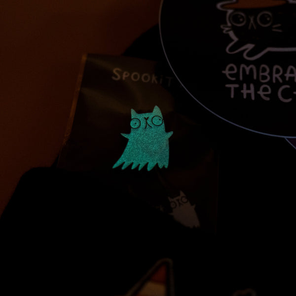 A close up of the glow in the dark Spookitty enamel pin from the Katie Abey Feline Spooky Box of Brilliance. The pin glows bright green in a dark setting, showing a ghost shaped cat with wide eyes. Part of Katie Abey’s quirky illustrated collection celebrating weird, fun and spooky vibes.