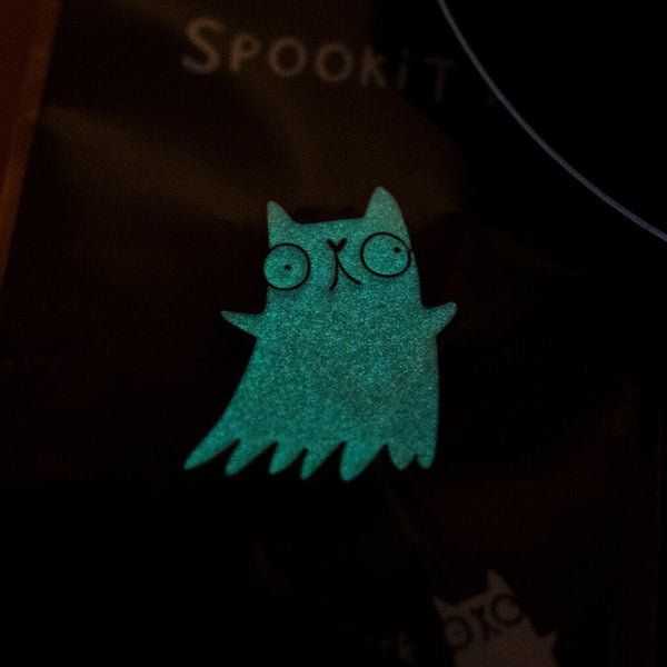 A Katie Abey Spookitty glow in the dark glitter enamel pin badge glowing bright green in the dark. The ghost cat shape is clearly visible with round eyes and wavy edges. A playful spooky themed enamel pin that glows at night and stands out on jackets, bags, and accessories.