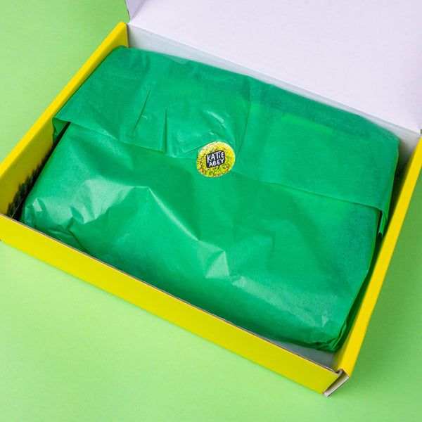 The Box of Brilliance - Subscription Boxes - Full Year Subscription on a green background. The yellow cardboard box is filled with green tissue paper with a mossy katie abey logo sticker sealing it shut.