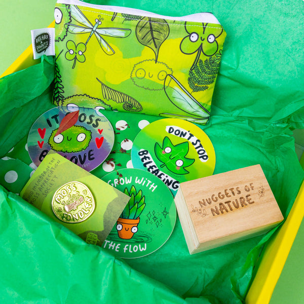 The Box of Brilliance - Subscription Boxes - One Time Purchase on a green background. The yellow cardboard box is filled with green tissue paper and the box contents featuring moss and leaf themed circle stickers, a green nature themed coin purse, a moss fondler pin and a wooden box with a crystal inside with text on the mini box reading 'nuggets of nature'.