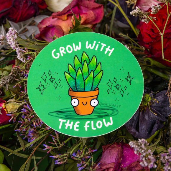 The Grow with the Flow Vinyl Sticker on purple, red, pink and green foliage. The green circular sticker has a smiling house plant with black sparkles and white text reading 'grow with the flow' surrounding.