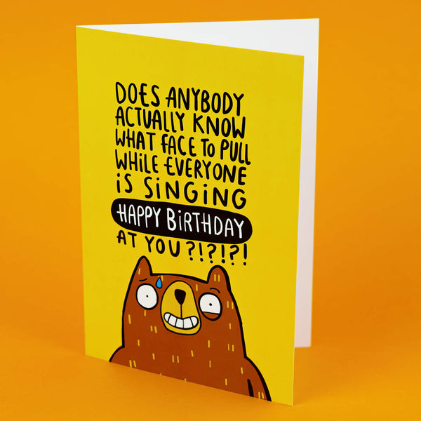 The Birthday Bear a6 Greeting Card designed and printed in the UK by Katie Abey. The card is stood upright on an orange background. The front cover is a yellow background with a stressed looking bear at the bottom smiling with a twitchy eye and bead of sweat whilst the text above them reads 'Does anybody actually know what face to pull while everyone is singing happy birthday at you?!?!?!'.