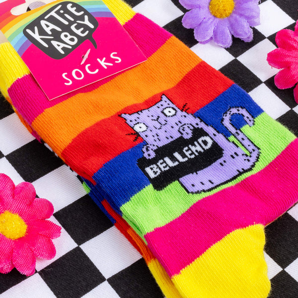 A pair of rainbow striped socks with a purple cat in the middle holding a sign saying bellend. They are on a checkerboard black and white background with flowers and ribbons