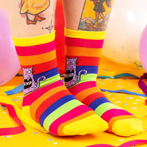 a model with tattooed legs wearing Katie Abey Bellend socks with purple cat holding a sign. They are rainbow striped and lovely and vibrant. The model is stood on a yellow floor with balloons, confetti and ribbons