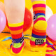 a model with tattooed legs wearing Katie Abey Bellend socks with purple cat holding a sign. They are rainbow striped and lovely and vibrant. The model is stood on a yellow floor with balloons, confetti and ribbons this shows the sole of the socks and the Katie Abey logo