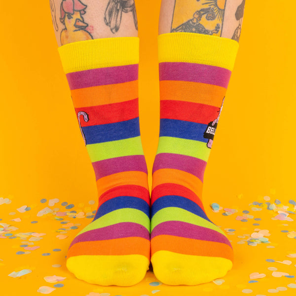 A front-facing shot of rainbow-striped socks with yellow cuffs and toes, featuring a purple cat design on the side. Confetti scattered on a yellow background adds a festive vibe.