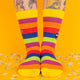 A front-facing shot of rainbow-striped socks with yellow cuffs and toes, featuring a purple cat design on the side. Confetti scattered on a yellow background adds a festive vibe.