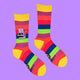 A pair of Katie Abey's rainbow striped socks laid flat, showing off a funky purple cat illustration holding a "Bellend" sign. These fun crew socks are an eye-catching mix of colour and personality, ideal for those embracing their weird side.