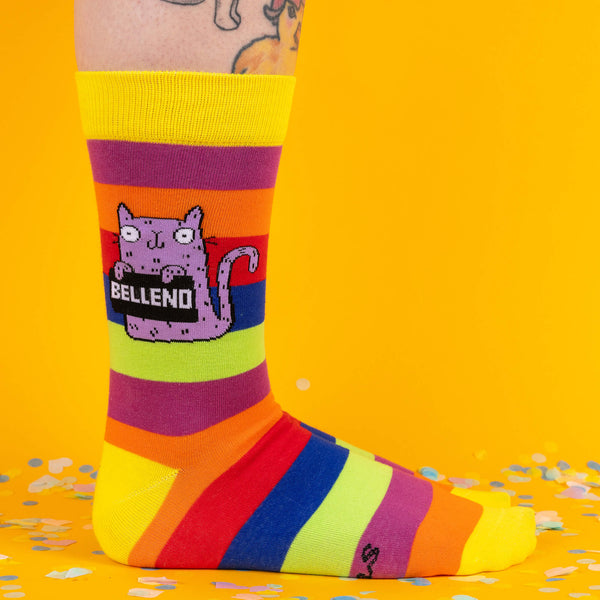 A side view of the socks showcasing the purple cat holding a sign that says Bellend, with vibrant rainbow stripes running throughout, set against a fun yellow backdrop.