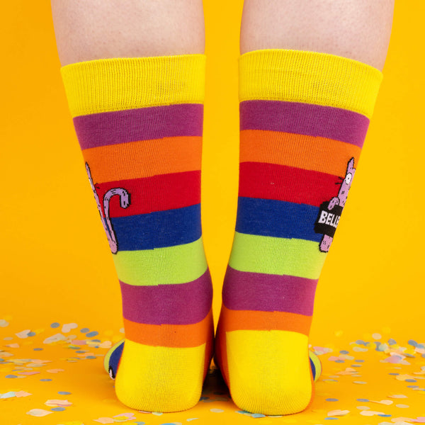 A back view of the socks, highlighting the bright rainbow stripes and the playful cat tail detail wrapping around the sides, against a cheerful yellow background with confetti.