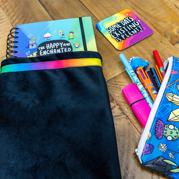A black oracle bag with a rainbow ribbon around the top with the happy and enchanted planner popping out the top. it has a coaster and pencil case in the shot on a wooden table