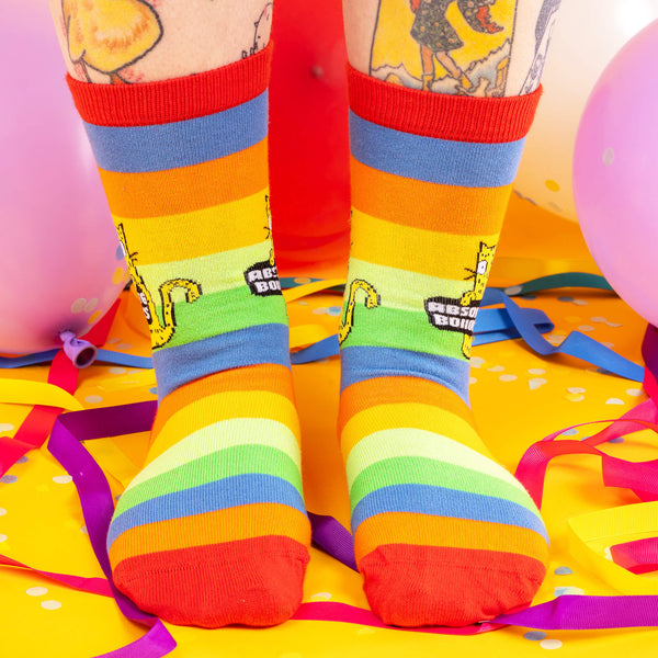 A model wearing Katie Abey socks with a yellow cat holding a sign saying absolute bollocks. They are rainbow striped and lovely and vibrant. The model is stood on a yellow floor with balloons, confetti and ribbons