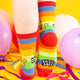 A model wearing Katie Abey socks with a yellow cat holding a sign saying absolute bollocks. They are rainbow striped and lovely and vibrant. The model is stood on a yellow floor with balloons, confetti and ribbons