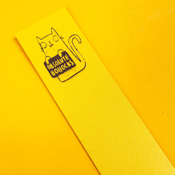 top of a yellow bookmark with an embossed cat illustration holding a sign saying absolute bollocks