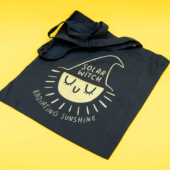 Black cotton tote bag by Katie Abey featuring a cute sun character wearing a witch hat with the text 'Solar Witch' and 'Radiating Sunshine' printed in light yellow. The bag is screen-printed for durability and photographed against a bright yellow background.