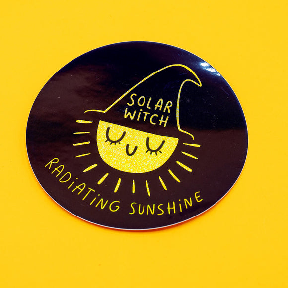A round vinyl sticker featuring a glittery yellow smiling sun wearing a witch hat with the words "Solar Witch" written on it. Around the sun, the phrase "Radiating Sunshine" is written in playful text. Designed by Katie Abey, an illustrator encouraging you to embrace your weirdness. Perfect for laptops, notebooks, mirrors, and water bottles.