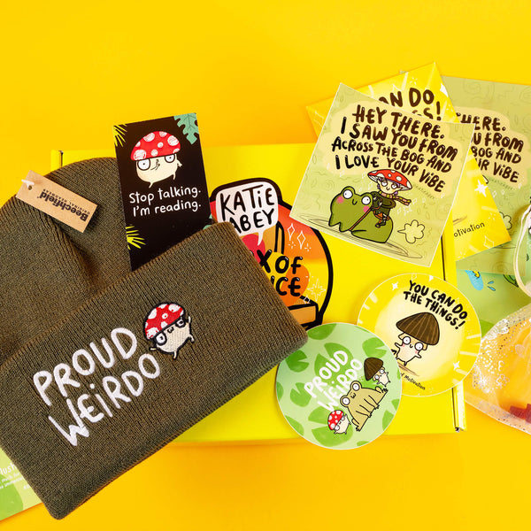 A flat lay of Katie Abey's "Proud Weirdo" box contents, including a green beanie with an embroidered mushroom character, quirky motivational stickers, and a yellow gift box. Texts like "You Can Do the Things!" and "I love your vibe" showcase fun, empowering designs perfect for embracing your weird.