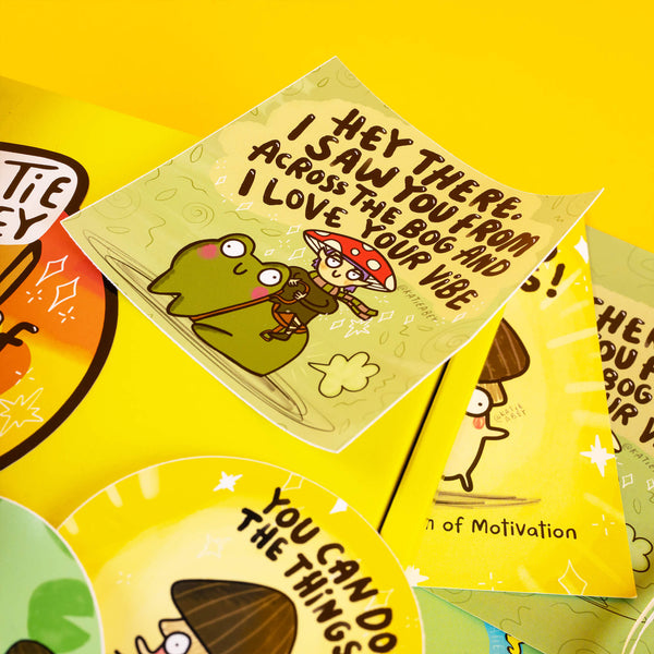 A yellow gift box from Katie Abey, showcasing a frog-themed card that says, "Hey there, I saw you from across the bog and I love your vibe," alongside motivational stickers with fun mushroom characters and vibrant colours. Celebrates embracing weirdness and spreading positivity with quirky illustrations.