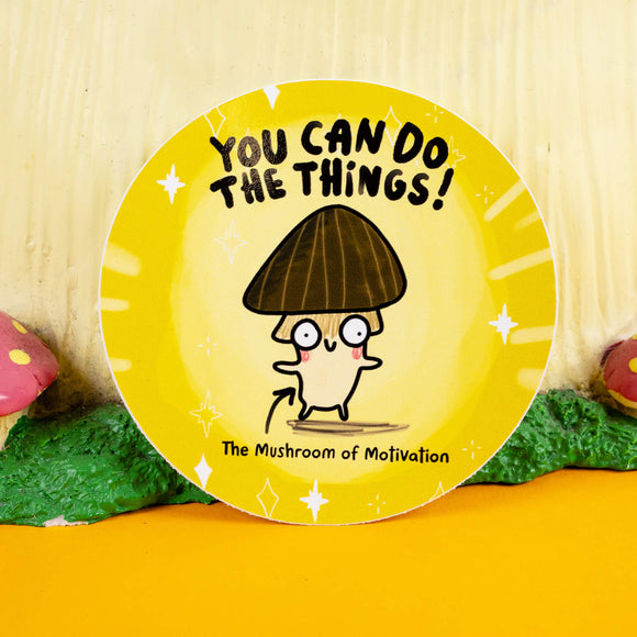 Quirky circular sticker by Katie Abey showcasing the "Mushroom of Motivation" illustration with a dark brown cap, rosy-cheeked face, and fun text that says "You Can Do The Things!" in bold black. The yellow background with sparkles exudes encouragement and charm, set against a whimsical woodland-inspired backdrop.