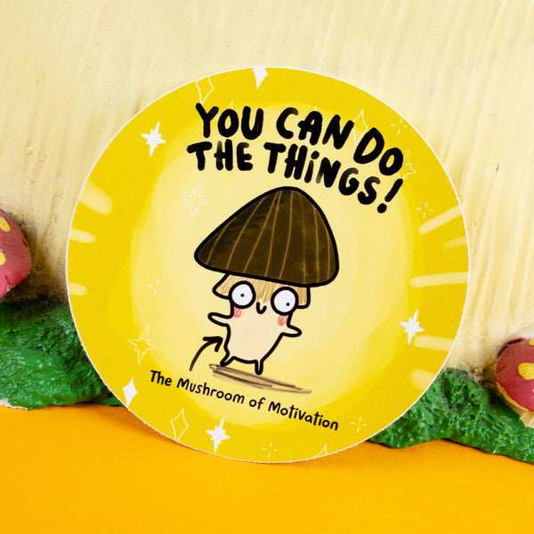 Circular motivational sticker featuring a whimsical mushroom with a dark brown cap, quirky eyes, and rosy cheeks. The text reads "You Can Do The Things!" in bold black lettering, with "The Mushroom of Motivation" written below. Designed by Katie Abey, this bright yellow sticker with sparkly accents inspires positivity and adds charm to any surface. Displayed on a woodland-themed setup with playful details.