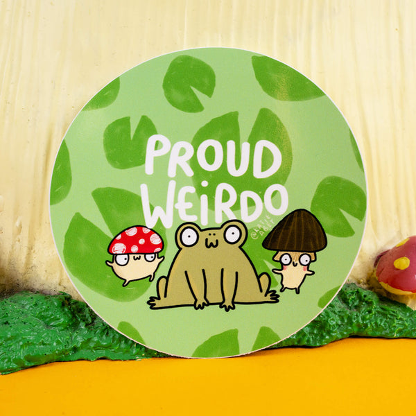 Circular sticker featuring a whimsical design by Katie Abey with the phrase "Proud Weirdo" in bold white letters. The illustration includes a quirky frog and two mushrooms (a red spotted cap and a dark brown cap) against a leafy green background. Katie Abey's small business celebrates individuality and embracing weirdness with this vibrant, playful design. The sticker is displayed in a fun, woodland-inspired setting.