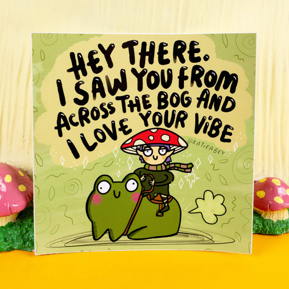 Quirky sticker design by Katie Abey featuring a mushroom elf riding a smiling frog alongside the text "Hey there. I saw you from across the bog and I love your vibe." The green background includes playful swirls and sparkly accents. Celebrating weirdness and good vibes, this sticker adds charm to any surface. Displayed on a natural woodland backdrop.