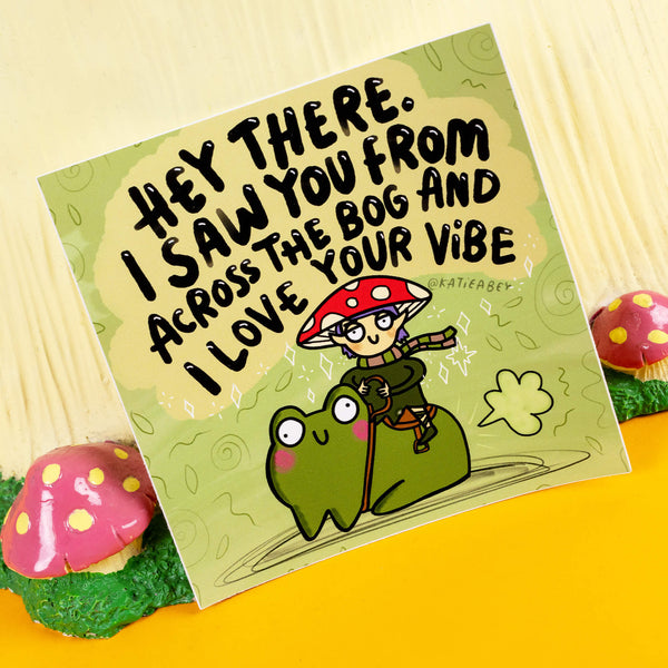 Square sticker by Katie Abey featuring a whimsical illustration of a red-spotted mushroom elf riding a happy frog, paired with the playful text "Hey there. I saw you from across the bog and I love your vibe." The green background is accented with swirls, sparkles, and leafy details. This quirky design celebrates individuality and spreading good vibes. Displayed in a fun woodland setting with toadstool props.