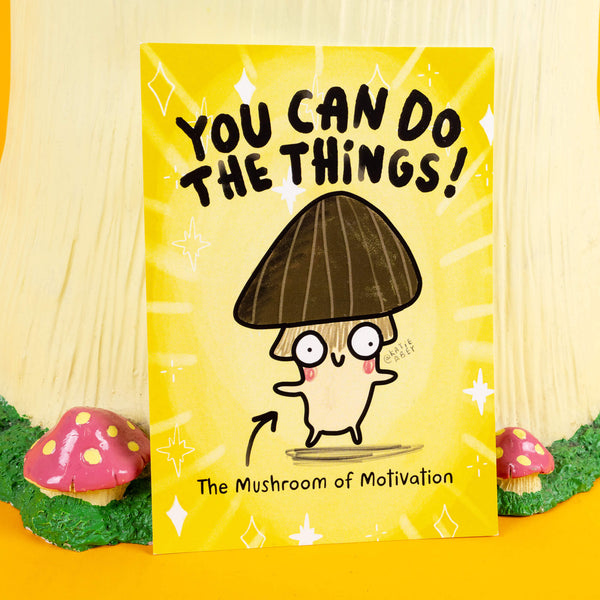 A bright yellow motivational print by Katie Abey featuring "The Mushroom of Motivation." The quirky illustration showcases a cute mushroom character with a brown cap, rosy cheeks, and wide eyes, paired with bold text reading "You Can Do The Things!" Sparkling accents and a fun vibe radiate positivity. This print, displayed in a whimsical woodland-themed setup with toadstool props, embodies Katie Abey's encouragement to embrace your unique self and achieve your goals.