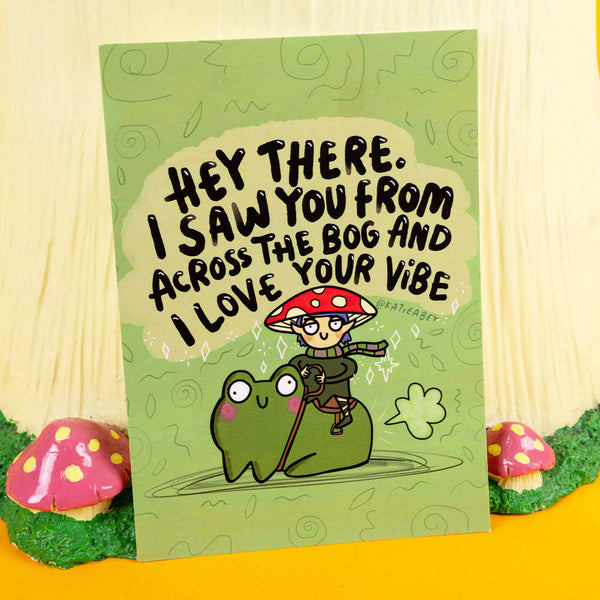 Katie Abey's quirky postcard design features a mushroom elf with glasses and a red-spotted cap riding a smiling frog, alongside the playful text "Hey there. I saw you from across the bog and I love your vibe." The green background, adorned with swirling patterns and leaves, enhances the whimsical vibe. A perfect way to spread positivity and celebrate weirdness.