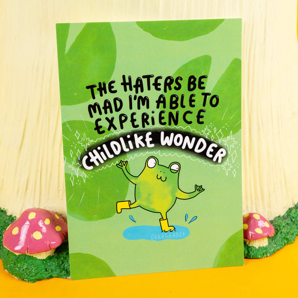 Katie Abey’s postcard showcases a fun-loving frog in bright yellow wellies joyfully splashing in a puddle, paired with the bold text, "The haters be mad I’m able to experience childlike wonder." Set against a leafy green background with sparkles, this design encourages embracing your inner child and spreading positivity. Ideal for quirky gifting or displaying as a daily reminder to find joy in the little things.