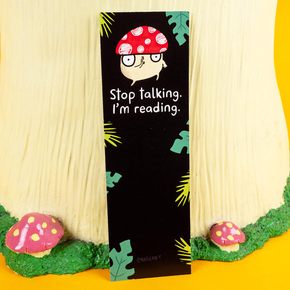 Katie Abey bookmark featuring a quirky mushroom character with a red-spotted cap and glasses, accompanied by the bold text "Stop talking. I’m reading." The design sits against a black background with tropical leaf accents, blending playful charm and bold attitude. Perfect for book lovers who enjoy a dose of humour with their reading adventures. Displayed in a whimsical woodland setup with toadstool props.