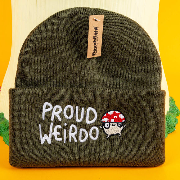 Olive green "Proud Weirdo" beanie with whimsical white text and a red-spotted mushroom character in glasses, showcasing Katie Abey's unique and quirky style. Small business Katie Abey inspires embracing your inner weirdness. Beanie displayed on a fun woodland-inspired setup.