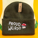 Olive green "Proud Weirdo" beanie with whimsical white text and a red-spotted mushroom character in glasses, showcasing Katie Abey's unique and quirky style. Small business Katie Abey inspires embracing your inner weirdness. Beanie displayed on a fun woodland-inspired setup.