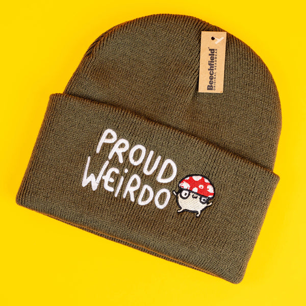 Olive green knitted beanie featuring "Proud Weirdo" text in playful white embroidery alongside an adorable mushroom character with a red spotted cap and glasses. Katie Abey's quirky, illustrated design encourages individuality and celebrates weirdness. Beanie shown flat with vibrant yellow background.