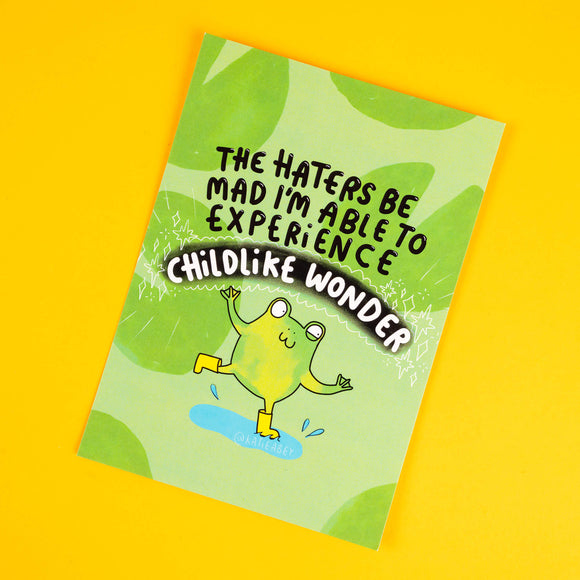 Katie Abey postcard featuring a vibrant green design with a cheerful frog in yellow wellies splashing in a puddle. The bold text reads, "The haters be mad I’m able to experience childlike wonder." This quirky and uplifting illustration celebrates embracing joy and individuality, surrounded by sparkles and playful leafy accents. Perfect for adding a touch of whimsy and positivity to your space or sending as a fun and encouraging gift.