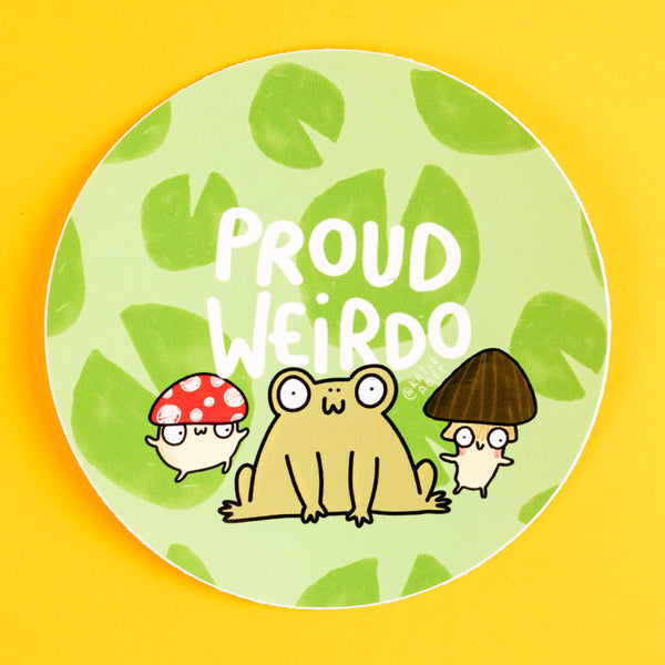 Quirky "Proud Weirdo" sticker by Katie Abey with a cheerful green background filled with leafy patterns. The design features a cute frog and two mushroom characters—one with a red spotted cap, the other with a dark brown cap—celebrating individuality. Displayed on a bright yellow backdrop, this circular sticker adds charm to any surface.