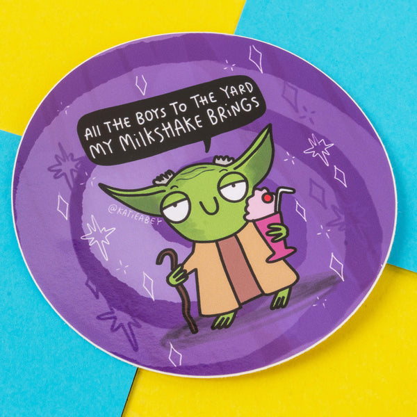 A vibrant, circular sticker featuring an illustration by Katie Abey, depicting a green alien-like character holding a pink milkshake and a walking stick. The character humorously says, "All the boys to the yard my milkshake brings" in a speech bubble. The sticker has a purple background with playful white star and diamond accents. Katie Abey is a UK-based illustrator and small business that celebrates embracing your weirdness and spreading joy through quirky designs.