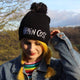 Brain cosy black knitted beanie bobble hat with grey pom pom and white embroidered lettering worn by Katie Abey stood amongst greenery. Designed by Katie Abey in the UK
