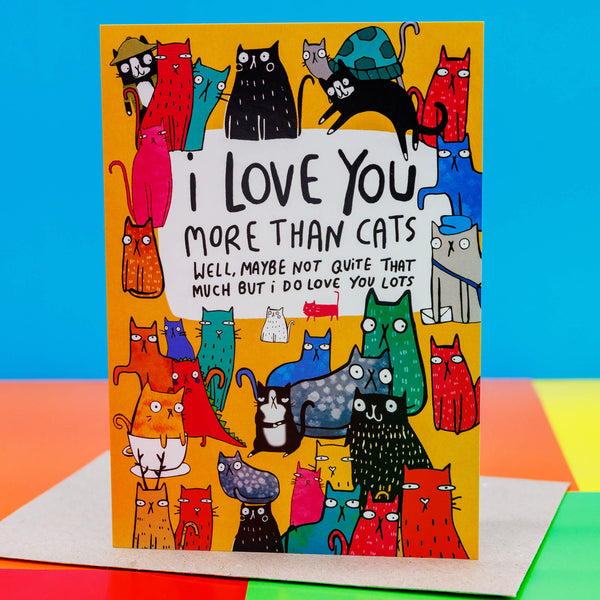 I love you more than cats a6 greeting card stood up on the brown envelope the card comes with, silver glittery paper and love hearts sweets on a white background. The card cover is a yellow background with various different coloured, patterned and sized cats with text in the top centre reading 'I love you more than cats. Well, maybe not quite that much but I do love you lots' Designed by Katie Abey in the UK.