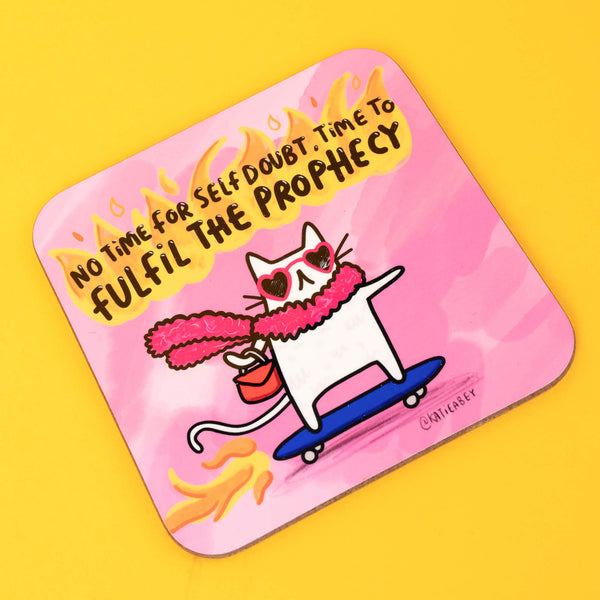 A pink coaster from Katie Abey featuring a quirky white cat wearing heart-shaped sunglasses and a fluffy pink scarf, skateboarding with flames in the background. The bold text reads, "No time for self-doubt, time to fulfil the prophecy," inspiring confidence and fun.