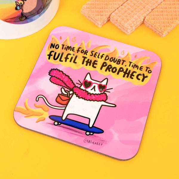 A fun pink coaster from Katie Abey, showcasing a white cat in heart-shaped sunglasses and a fluffy pink scarf on a skateboard with flames. The text, "No time for self-doubt, time to fulfil the prophecy," adds a touch of playful motivation and charm.