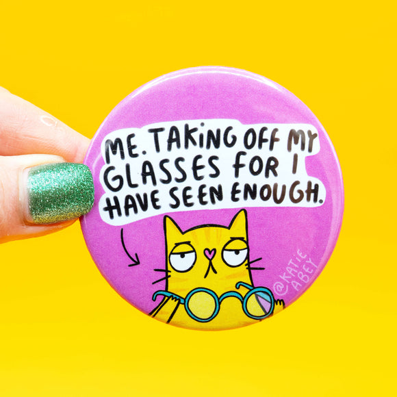 Close-up of a hand holding a Katie Abey badge that reads “Me. Taking off my glasses for I have seen enough.” The background is pink and the cat has an unimpressed expression, holding a pair of turquoise glasses.