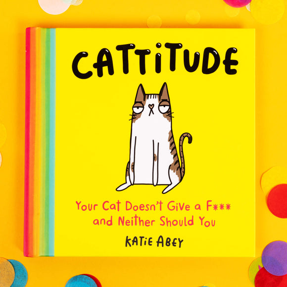 The front cover of the Cattitude Book illustrated by Katie Abey. It is bright yellow with a rainbow spine and an illustration of a tabby cat looking unamused with text above it that reads CATTITUDE. Underneath the cat there is text that reads Your Cat Doesn't Give a F*** and Neither Should You. The book is on a yellow background amongst coloured confetti.