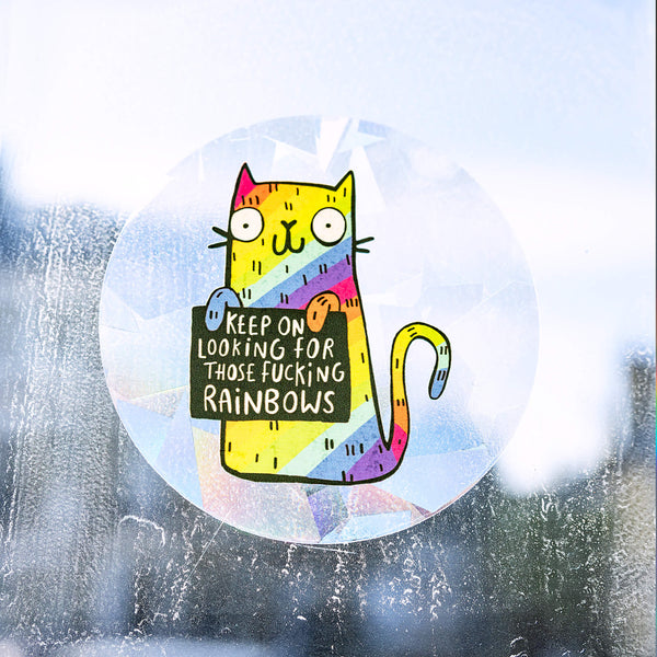 A see through rainbow making sun catching sticker that has a rainbow cat holding a sign saying keep on looking for those fucking rainbows